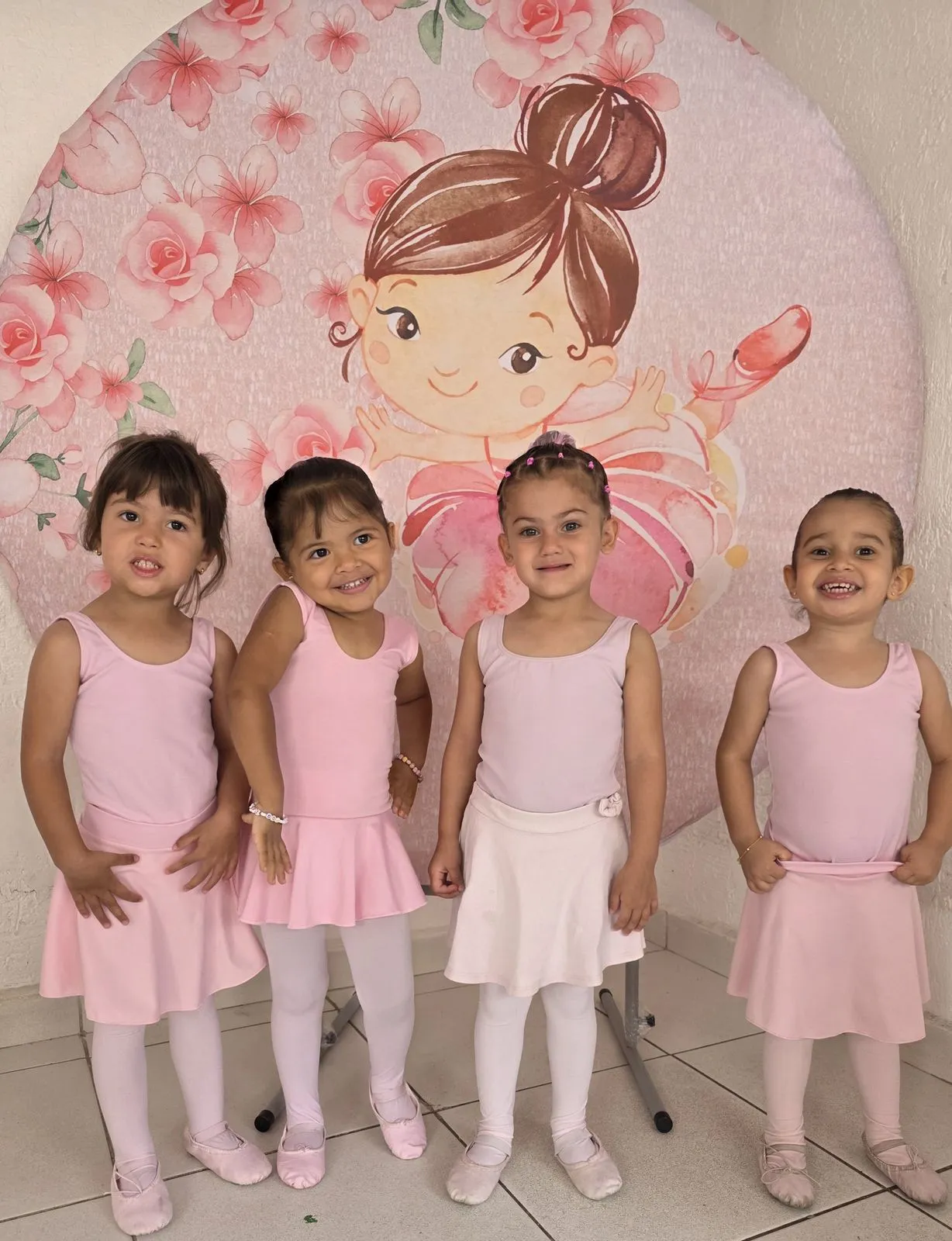 Quatro alunas da aula de balé em pé, de collant e saia rosa, diante de um painel com ilustração de fada