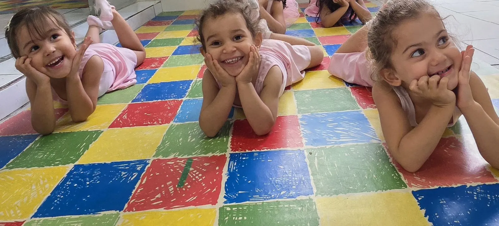 Duas alunas da aula de balé deitadas no tatame colorido, de collant rosa, olhando para a câmera